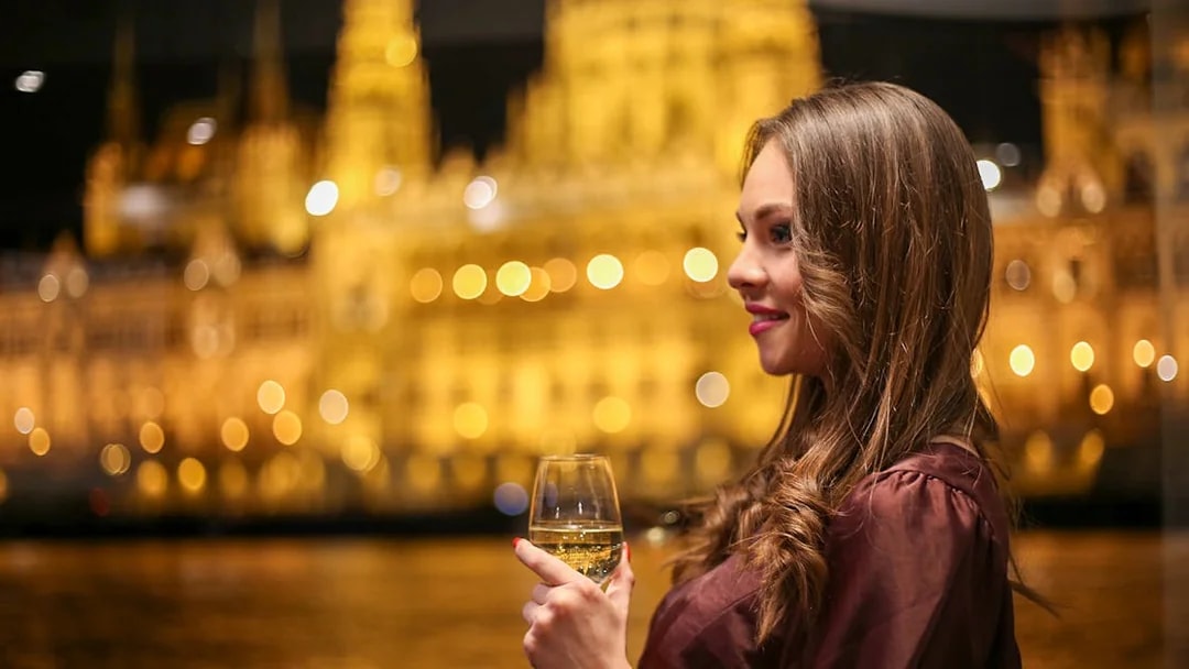 Woman enjoying a drink on Budapest dinner cruise with illuminated cityscape.
