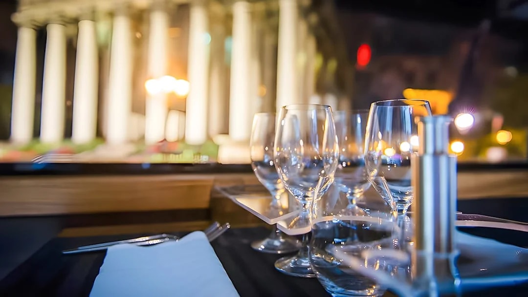 Cutlery and wine glasses on a table inside Bustronome Paris with blurred city lights.