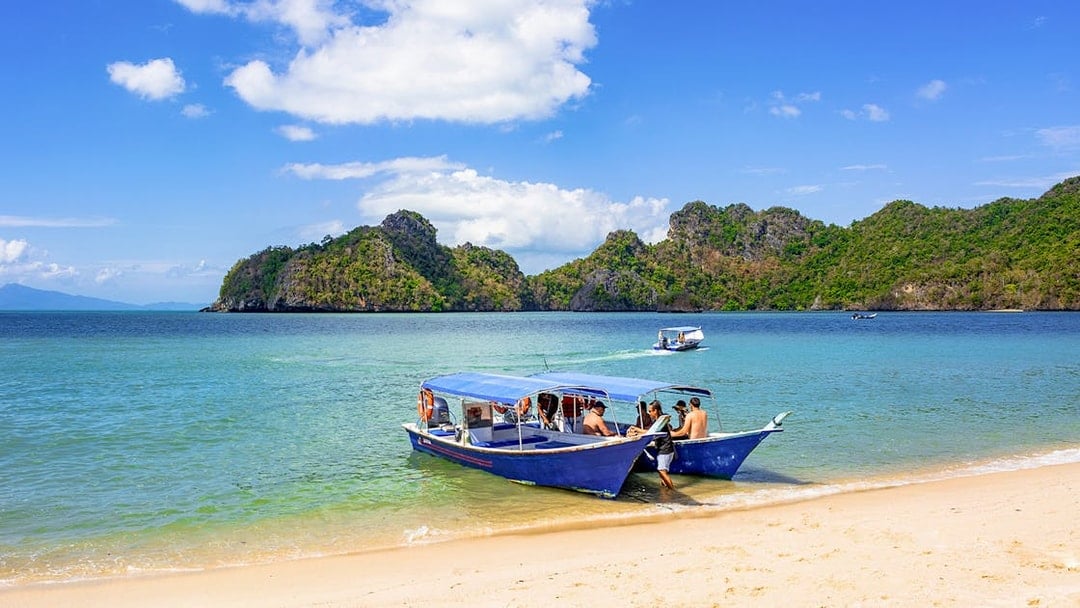 Boat docked on Langkawi beach with tourists, lush hills in background, part of Mangrove Discovery Adventure Tour.