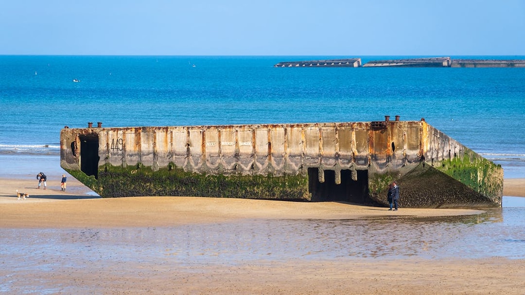 Remains of a D-Day landing craft on the beach in Caen, France, with visitors exploring nearby.
