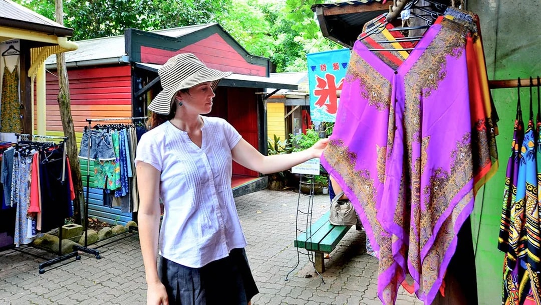 Woman shopping at Kuranda market, examining a colorful garment during the Full-Day Tour of Kuranda.