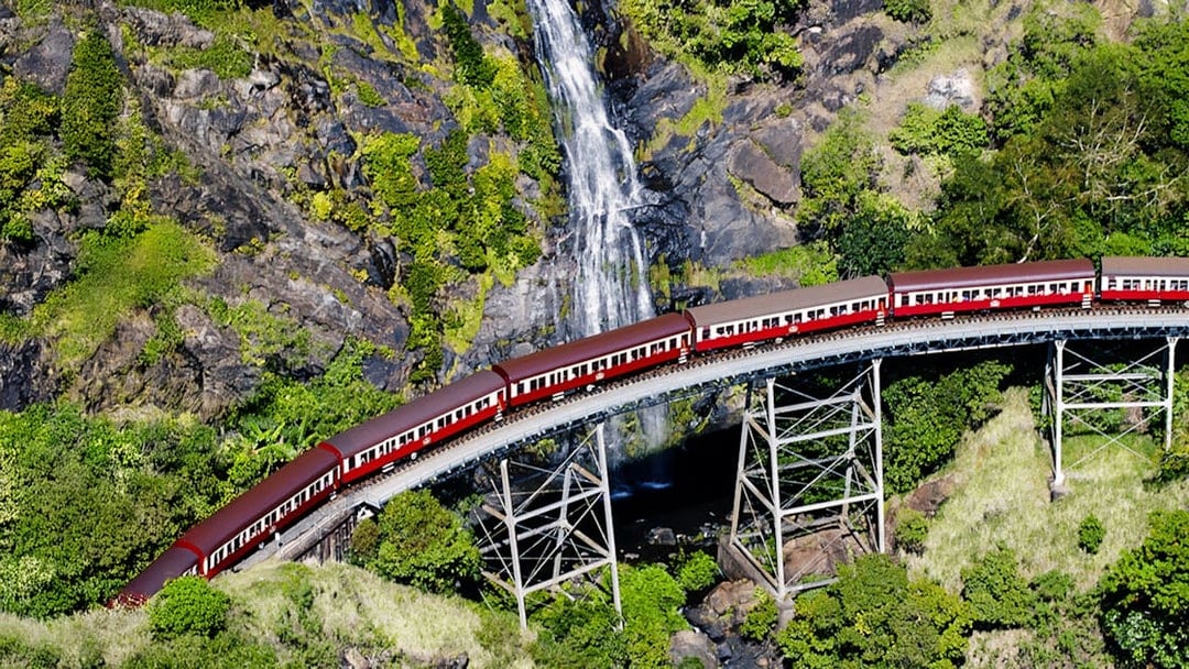 Aerial view of Kuranda Scenic Railway crossing a bridge with a waterfall in the background.