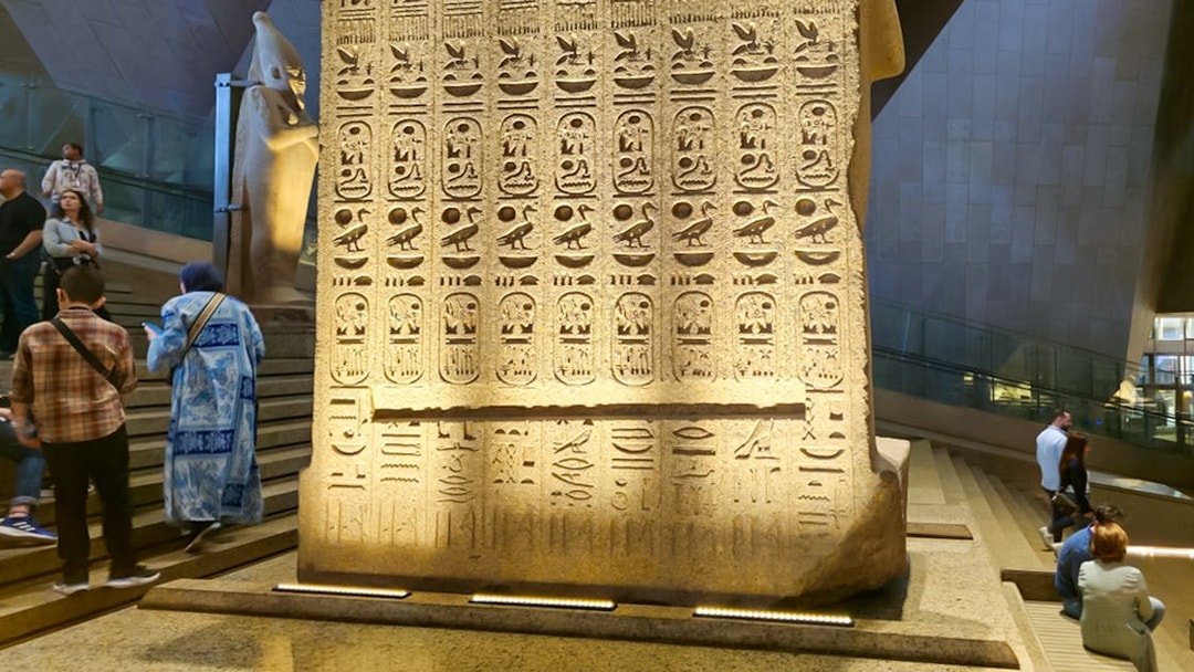 Hieroglyphs on stone slab at Grand Egyptian Museum, Cairo, with visitors exploring the exhibit.
