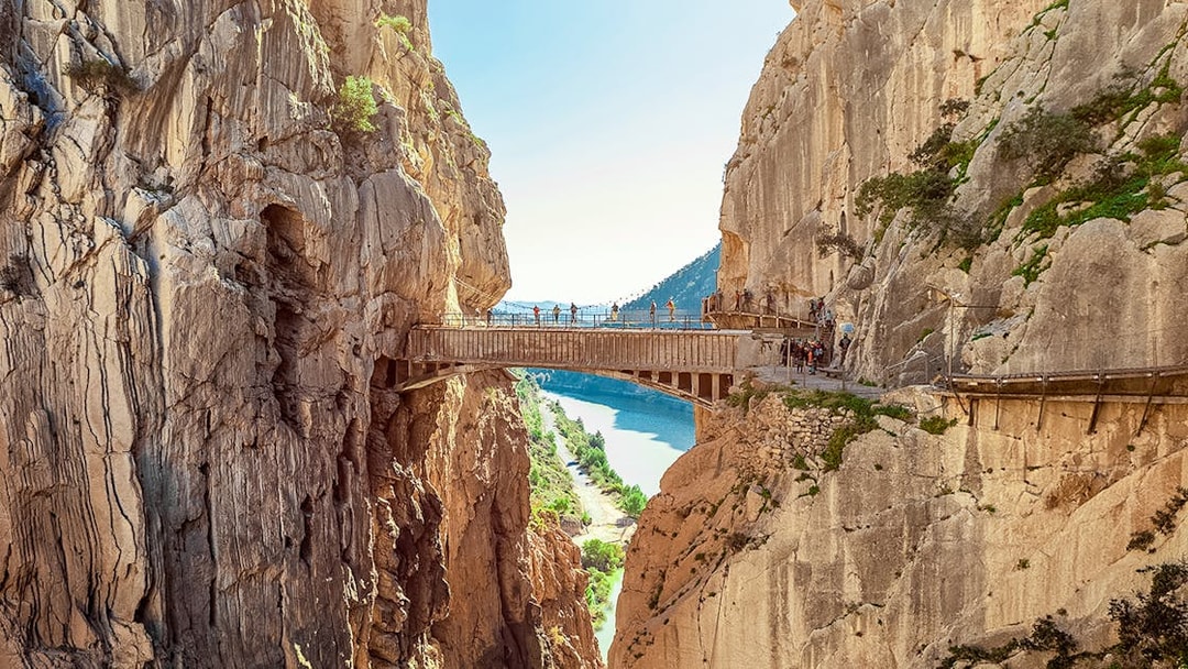 Hikers crossing a narrow bridge in Caminito del Rey, Spain, with cliffs and river below.