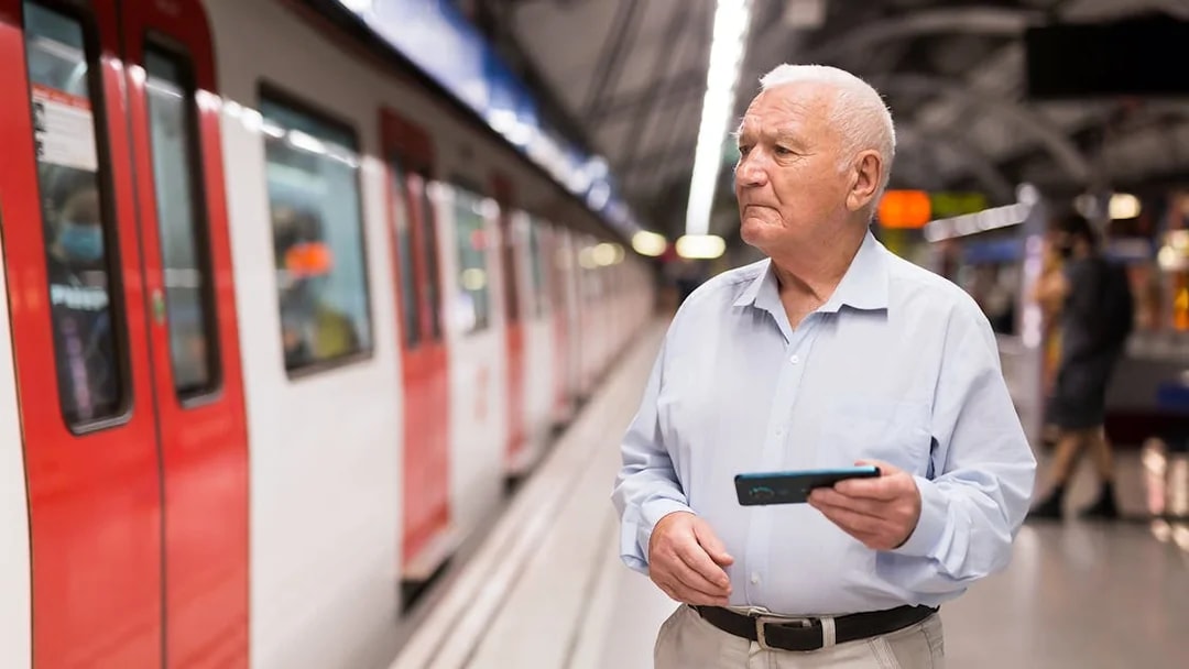 Elderly man holding a phone at a train station platform.