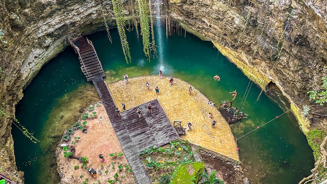 Tourists swimming in a natural Cenote pool surrounded by rocky cliffs and greenery.