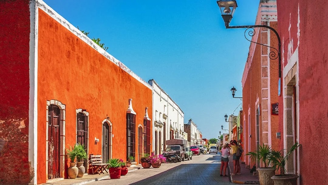 Colorful colonial buildings line a street in Valladolid, Mexico, with people walking and cars parked.