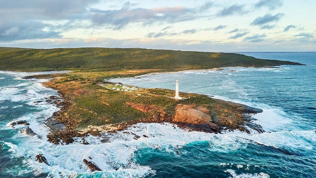 Aerial view of Cape Leeuwin Lighthouse on rocky coastal headland, surrounded by ocean waves.