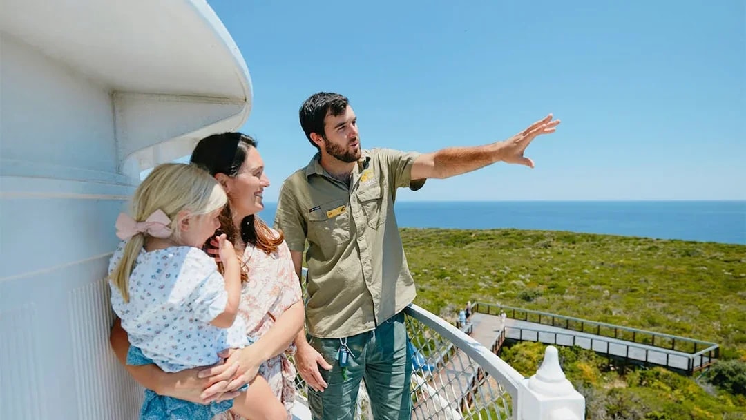 Tour guide points to ocean view from Cape Naturaliste Lighthouse balcony with visitors.