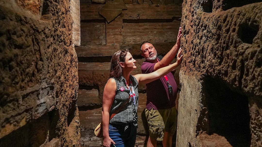Tourists exploring narrow passage in Roman Catacombs, touching ancient walls.