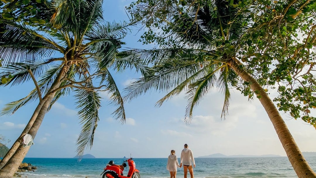 Couple on Langkawi beach with red motorbike under palm trees.