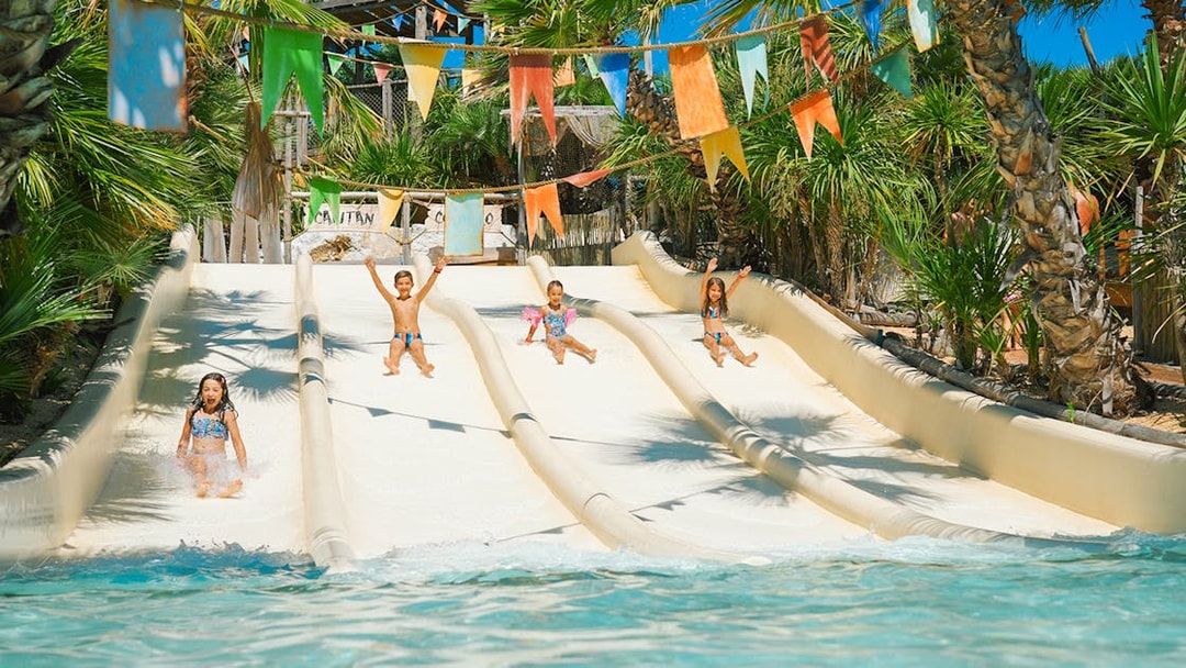 Kids sliding down water slides at Caribe Bay, surrounded by palm trees and colorful flags.