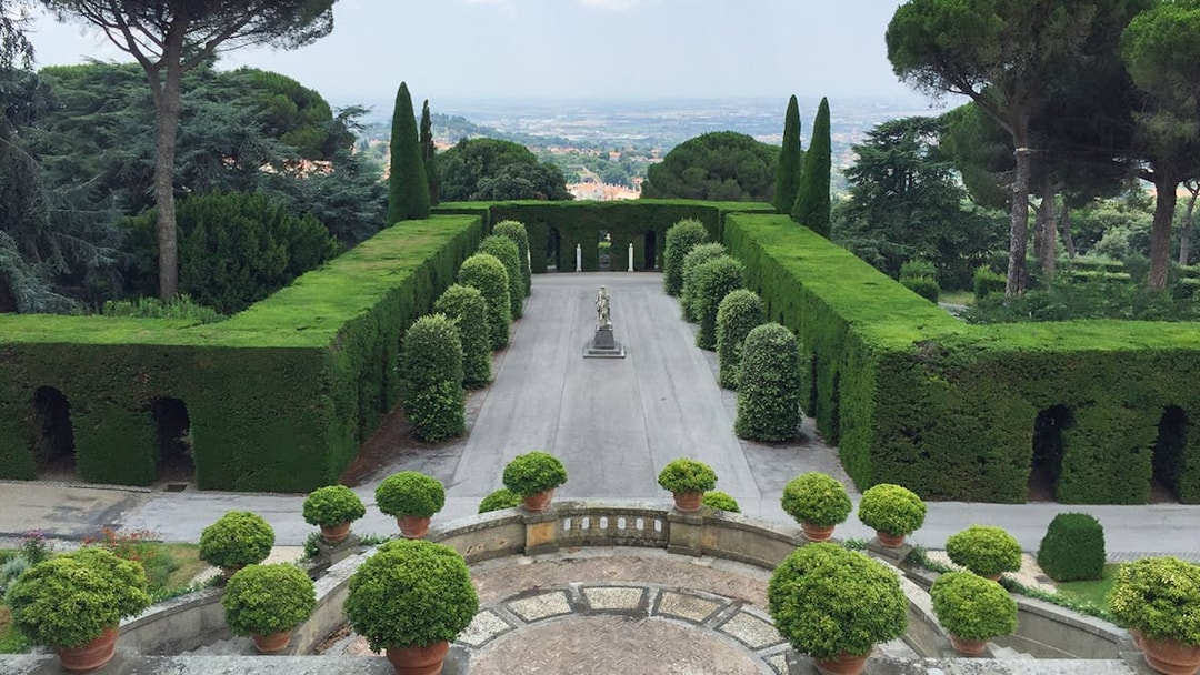 Castle Gandolfo garden with manicured hedges and central statue, overlooking scenic landscape.