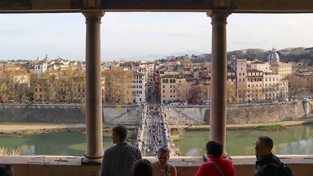 People walking on St. Angelo Bridge with a view of Rome's cityscape from Castel Sant'Angelo.