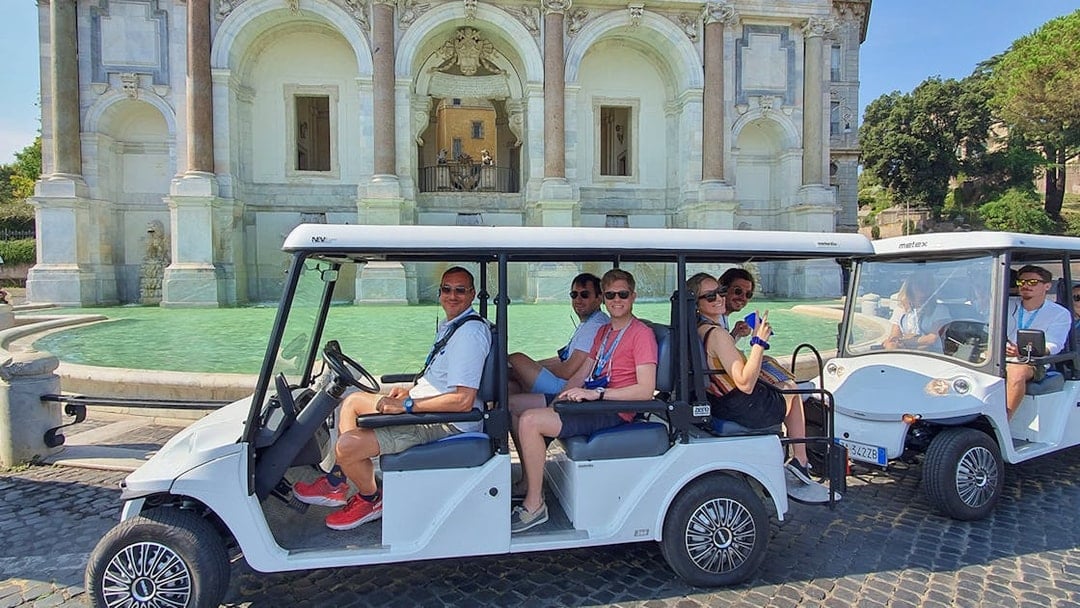 Golf cart with people touring near a historic fountain.