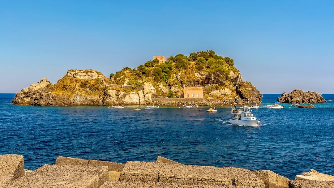 Snorkeling boats near Isole Ciclopi, Catania, with rocky island and clear blue sea.