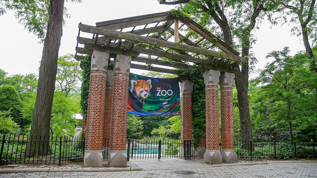 Entrance gate to a zoo with a banner featuring a red panda, surrounded by trees.