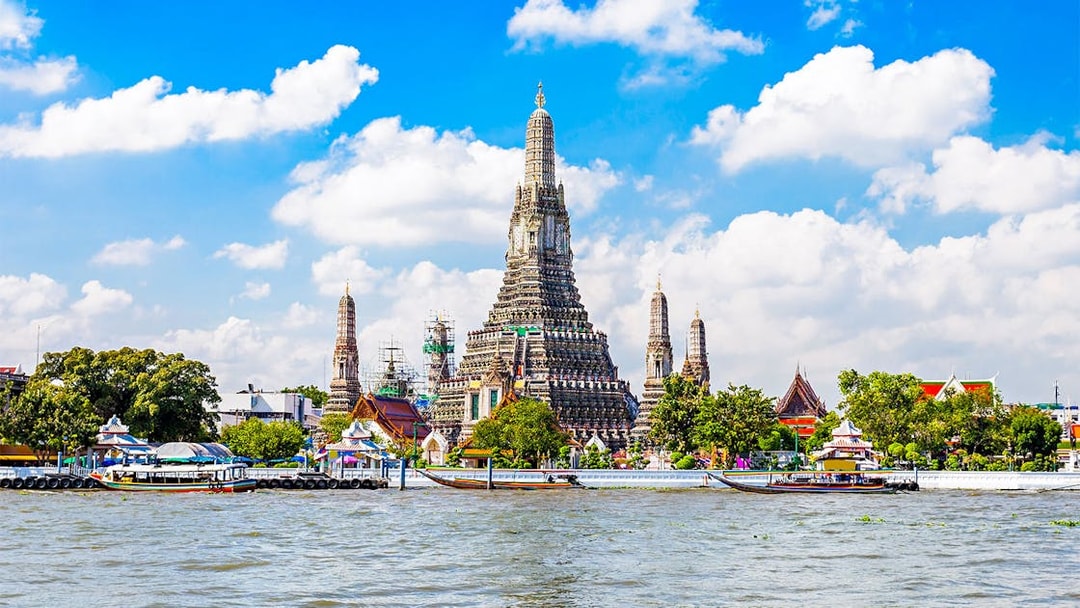 Wat Arun temple along Chao Phraya River in Bangkok with boats passing by.