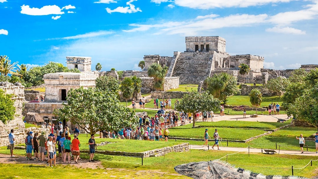 Iguana on Tulum ruins with tourists exploring ancient site in Mexico.