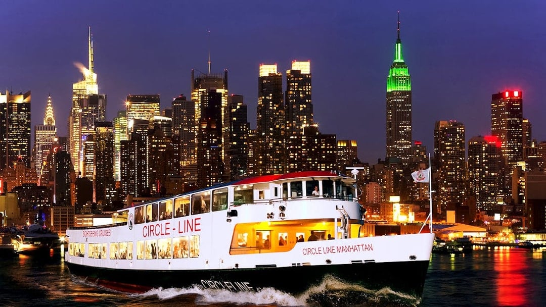 Tourists on Circle Line cruise with New York skyline illuminated at dusk.