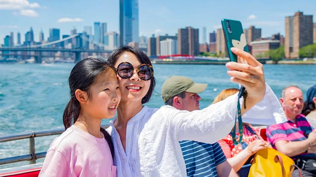 Tourists taking a selfie on a boat with New York City skyline and Brooklyn Bridge in the background.