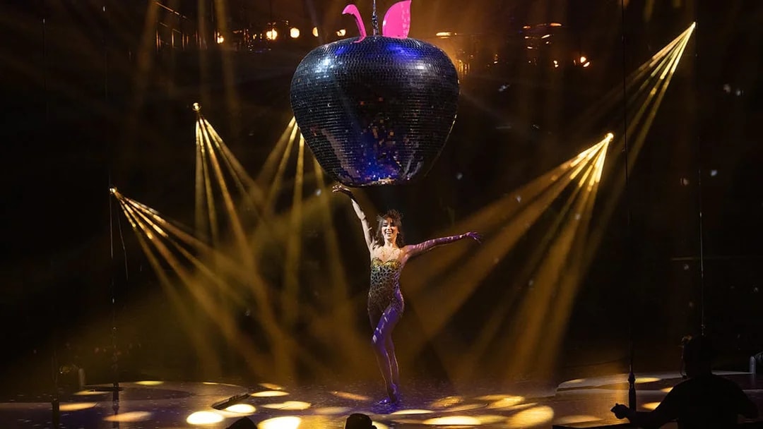Performer on stage with large disco apple prop in Mad Apple show.