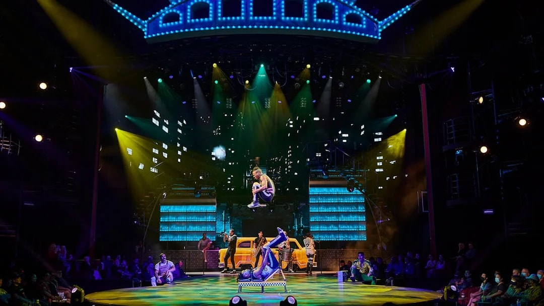 Acrobats performing on stage at Mad Apple show with vibrant cityscape backdrop.