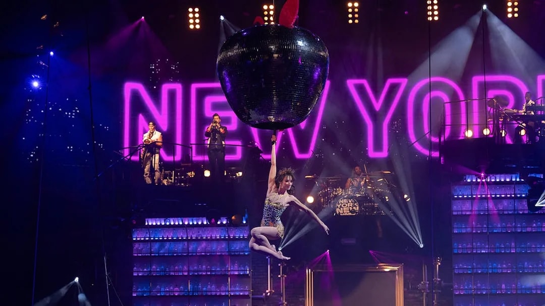 Performer hanging from a giant apple with "New York" sign in the background at Mad Apple show.