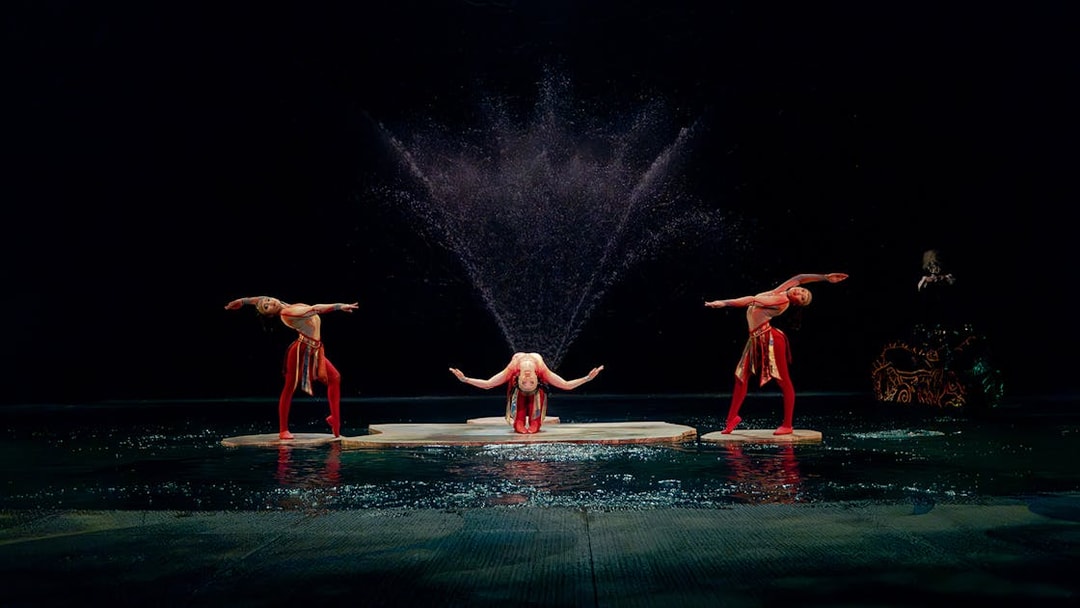 Performers in a water-themed acrobatic show with fountains in the background.