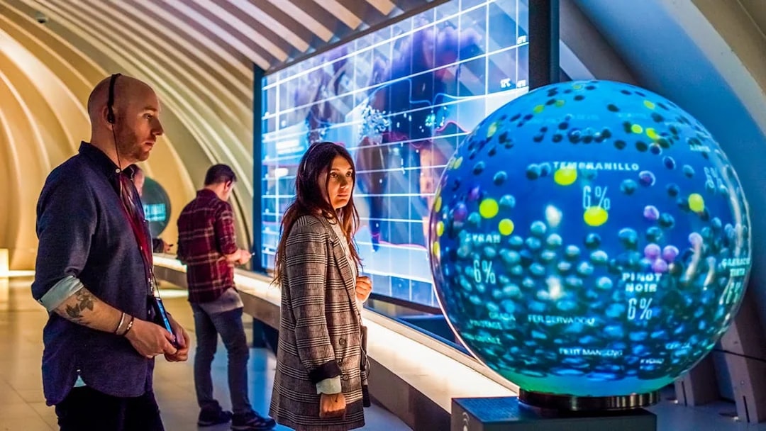 Tourists at Cité du Vin museum in Bordeaux using audio guides near interactive wine exhibit.