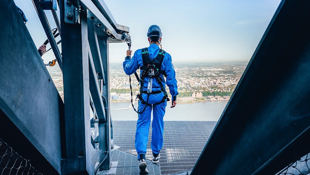 Person in safety gear walking on a high platform overlooking a city and river.