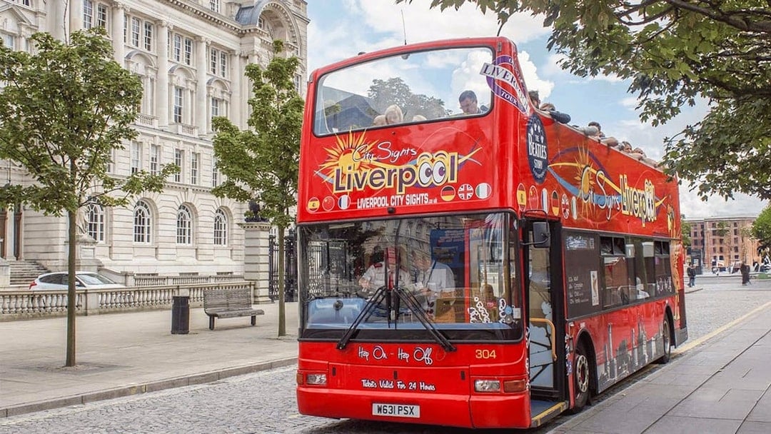 Red double-decker bus on Liverpool Hop On Hop Off City Tour near historic building.