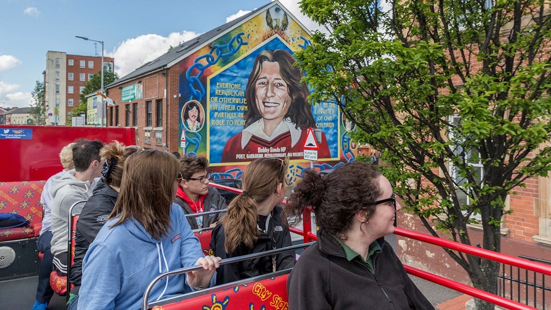 Tourists on Belfast hop-on-hop-off bus viewing mural of Bobby Sands on city street.