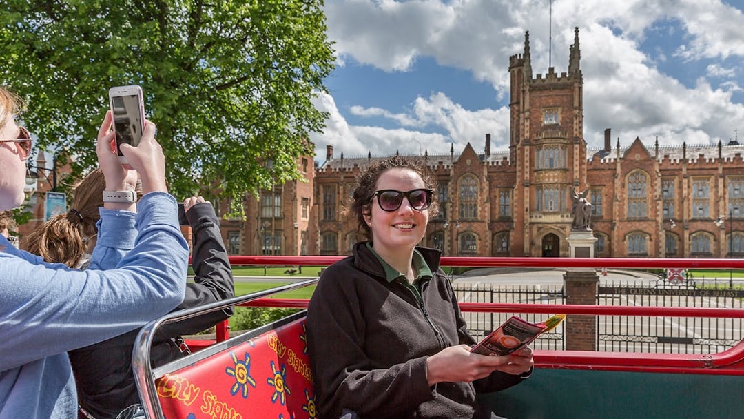 Hop-on hop-off bus near Queen's University Belfast with tourists taking photos.