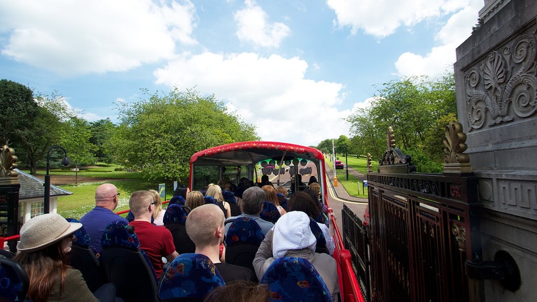 Open-top bus with tourists on Belfast Hop-On Hop-Off Tour passing through a park.