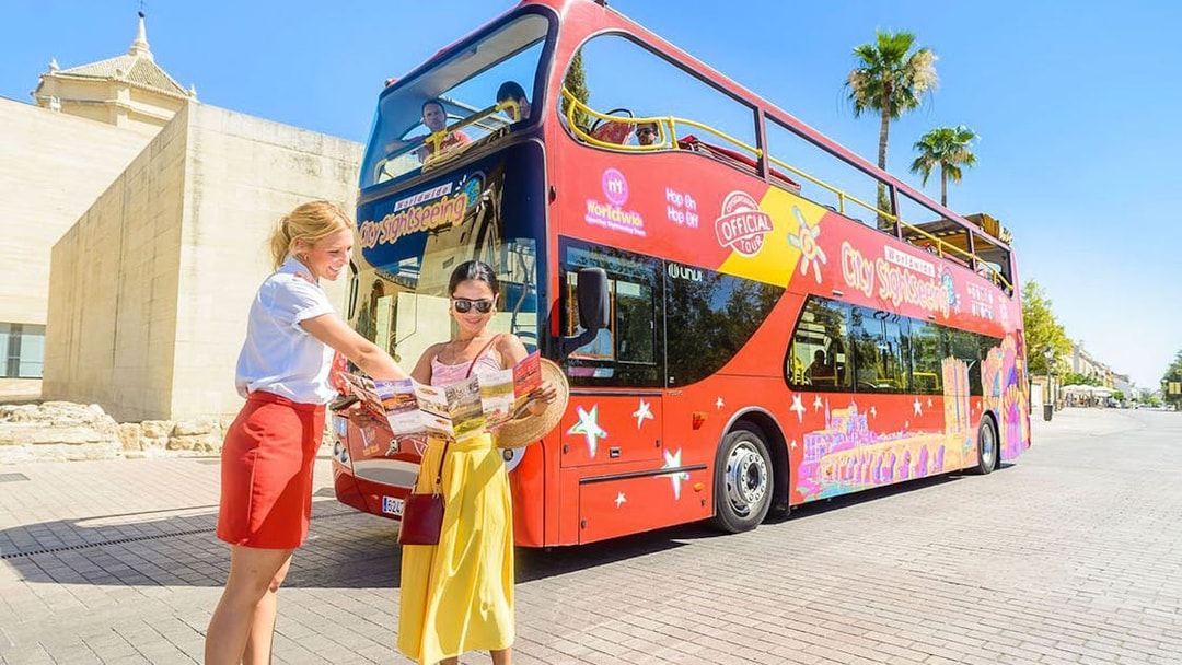 Woman with a passenger in front of a red hop-on hop-off sightseeing bus holding a map.