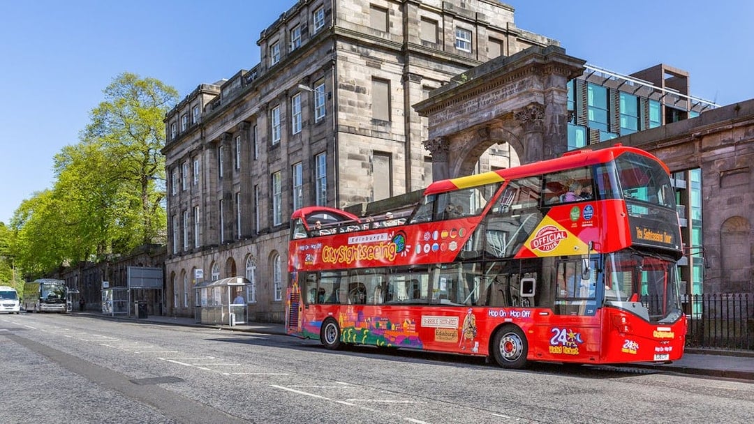 Red double-decker bus on Edinburgh Hop-On Hop-Off Tour near historic stone building.