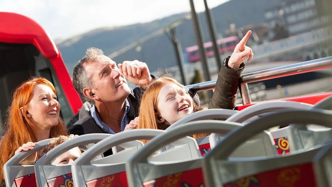 Family enjoying Edinburgh Hop-On Hop-Off bus tour, pointing at city sights.