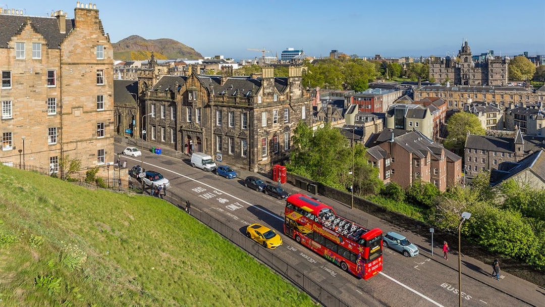 Hop-On Hop-Off bus on a street in Edinburgh with historic buildings and Arthur's Seat in the background.