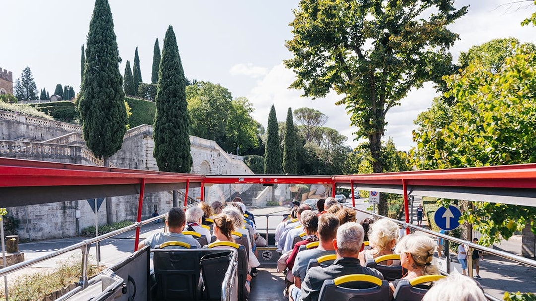 Open-top bus with tourists on Florence Hop-On Hop-Off tour passing historic stone wall and cypress trees.