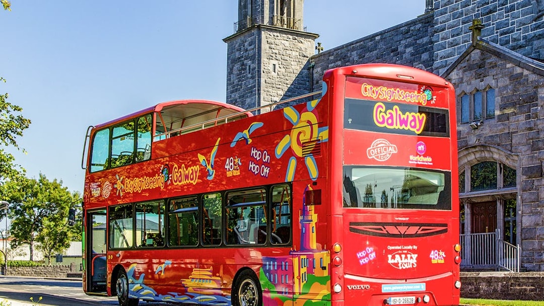 Red double-decker bus for Galway Hop-On-Hop-Off Tour near historic stone building.