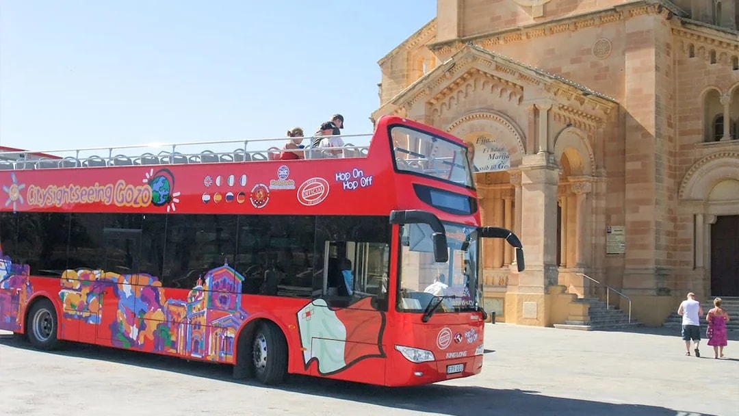 Red double-decker bus for Gozo Hop On Hop Off tour near a historic church.