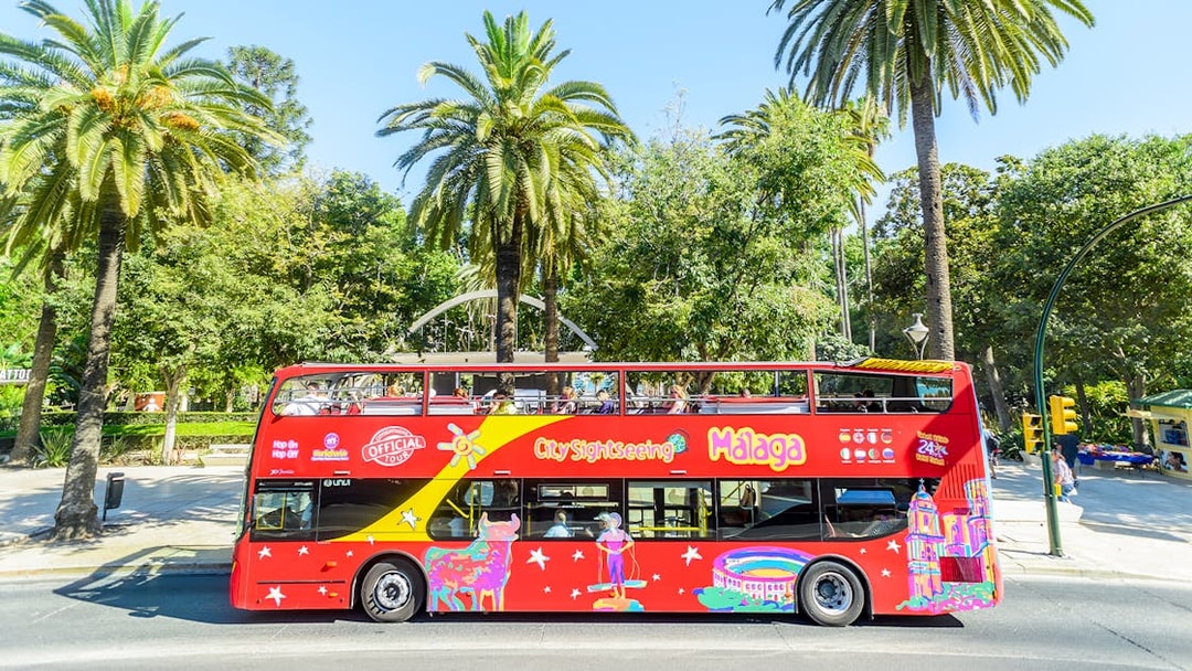 Red double-decker bus for Malaga hop-on hop-off tour near palm trees.