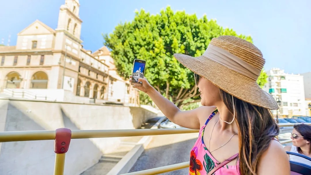 Woman taking photo from open-top bus on Malaga city tour.