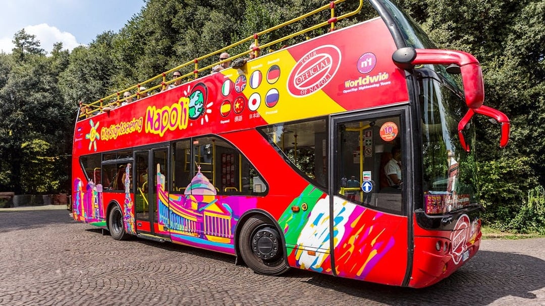 Red double-decker sightseeing bus in Naples with colorful designs and international flags.