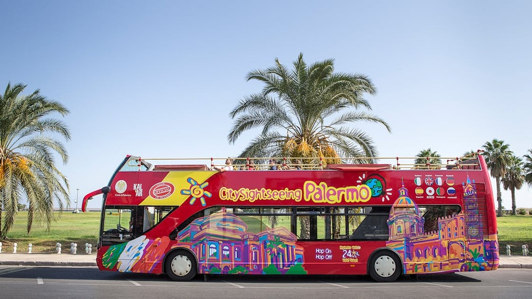 Palermo hop-on hop-off bus with colorful cityscape design parked by palm trees.