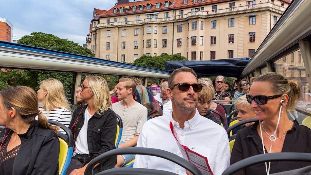 Open-top bus tour with passengers in Stockholm, historic building in background.