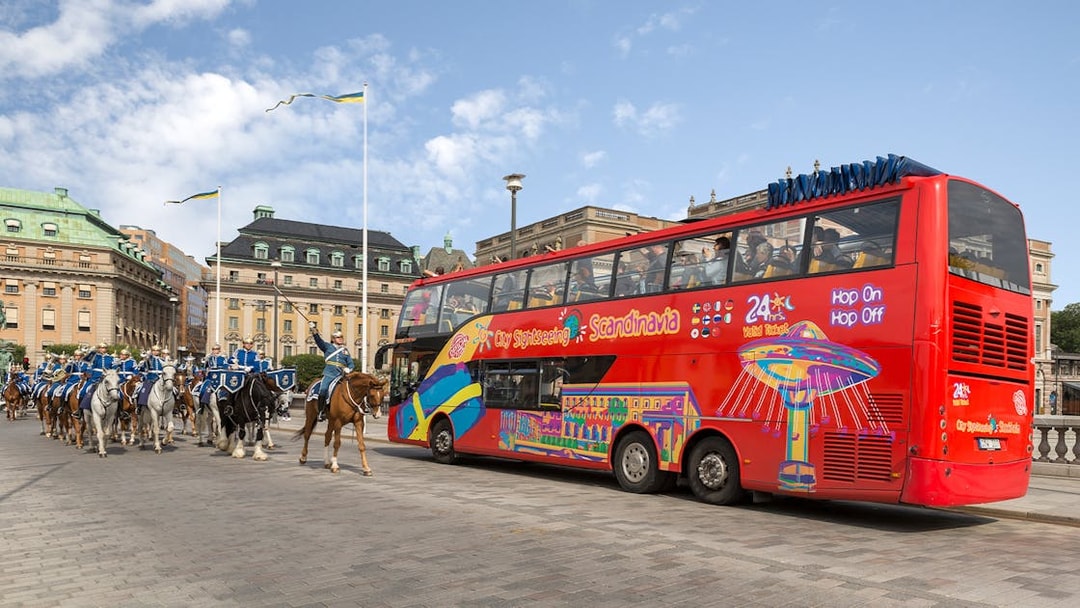 Red double-decker bus on a city sightseeing tour in Stockholm, passing a mounted royal guard parade.