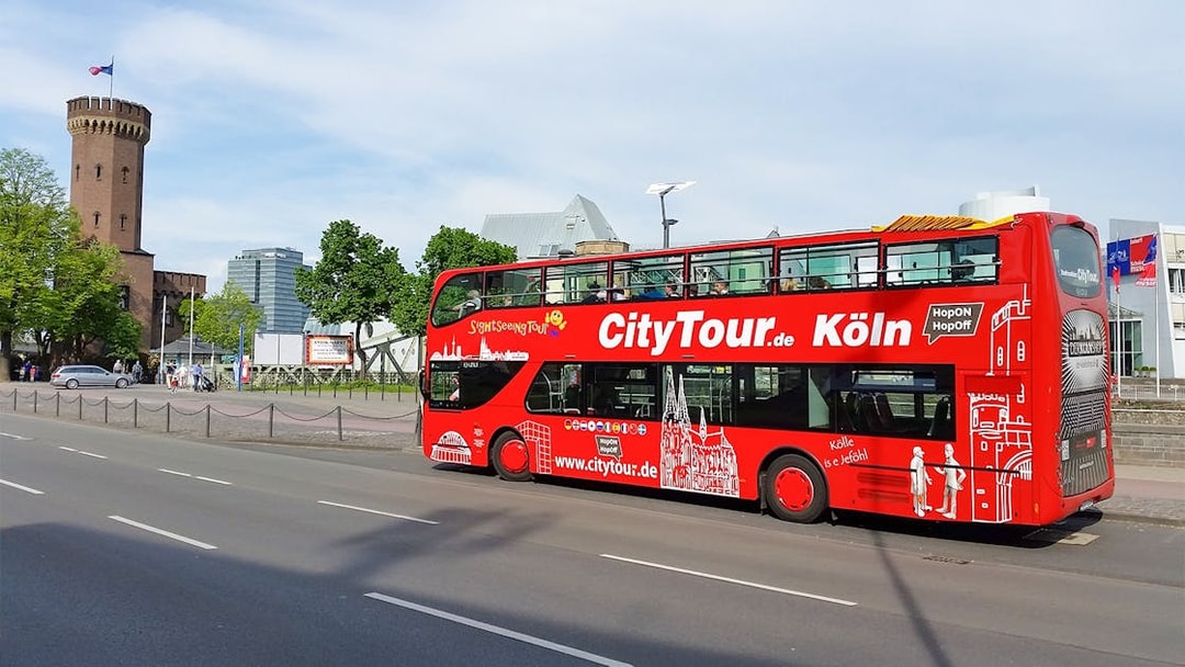 Red double-decker Hop on Hop off tour bus in Cologne near historic tower.