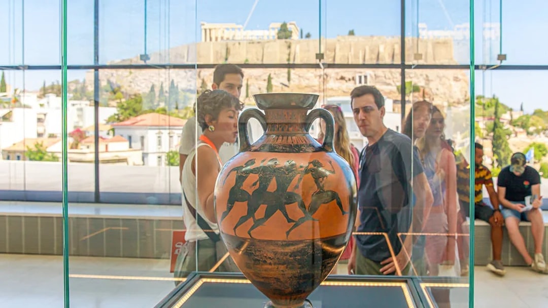 Visitors viewing ancient Greek vase at Athens Acropolis Museum with Parthenon in background.